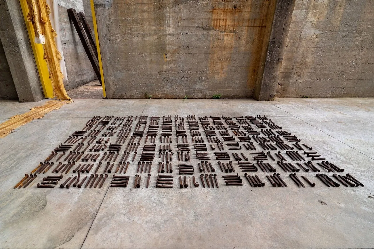 A concrete warehouse structure hosts a floor work: a grid of large, rusted metal spikes arranged in crosshatched sqaures.