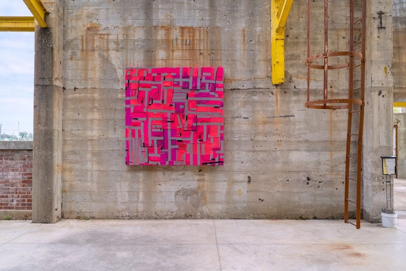 An open-aired concrete warehouse structure features a large, square wall work. The work appears to be covered with pink fabric with disorganized, patchworked lines of grey.