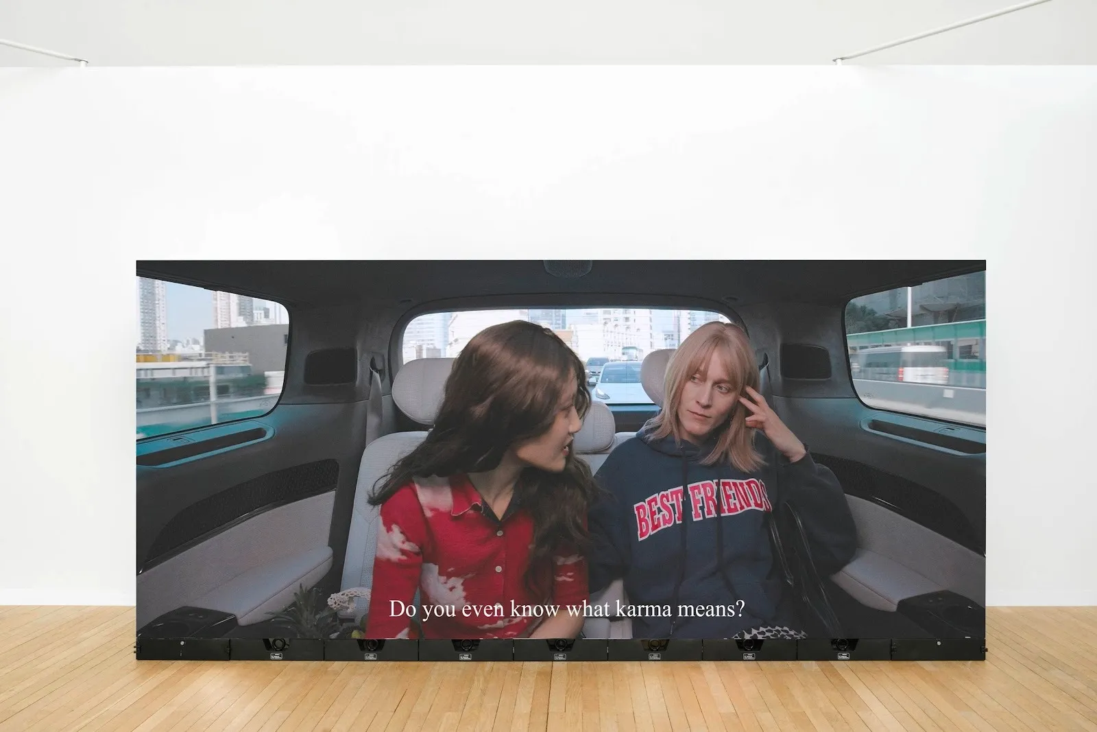 In a white room, a large LED screen features a video of two people talking to each other in a car. One speaks to the other, the subtitles reading, "Do you even know what karma means?"