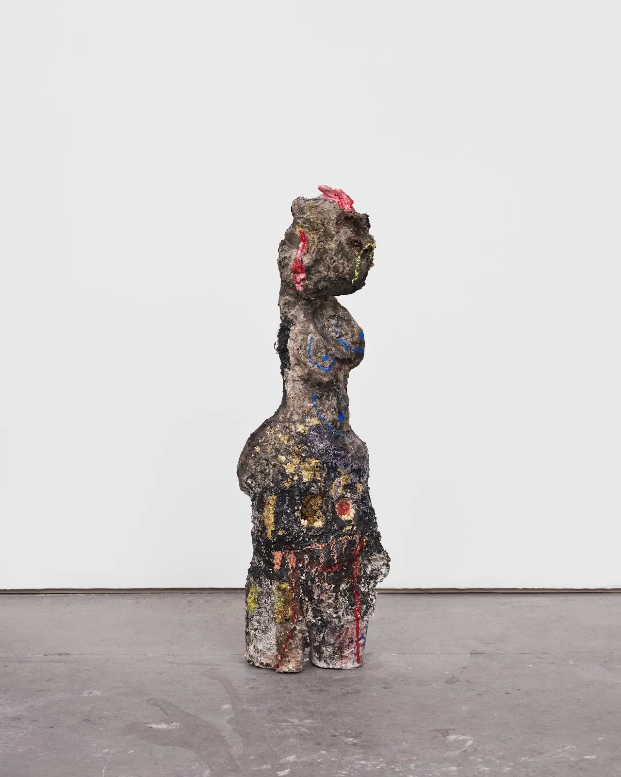 A heavily textured sculpture abstractly resembles a female body. The surface of the sculpture concrete-like and strewn with bright lines of pigment. The face is obscured.