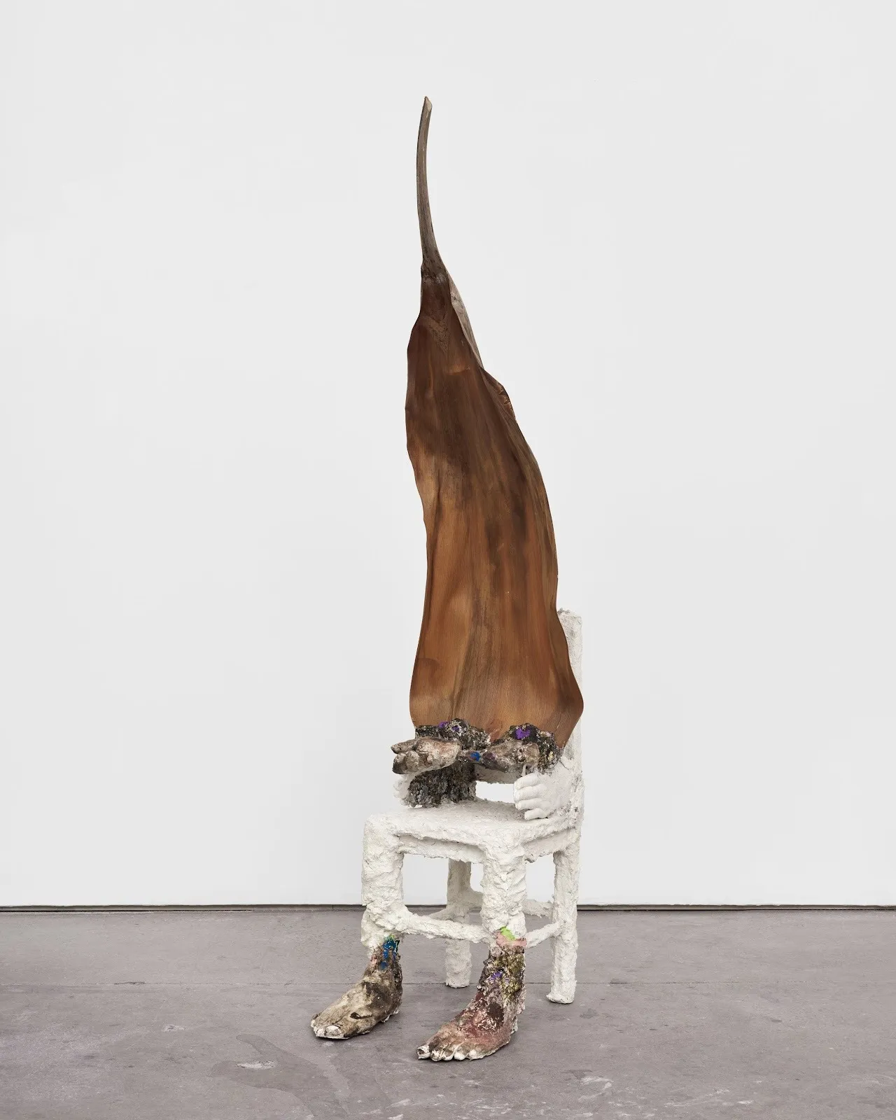 A textured white chair has two front legs that turn into lumpy human feet, while the armrests turn into outstretched hands. The back of the chair turns into a giant, brown pointed leaf shape.