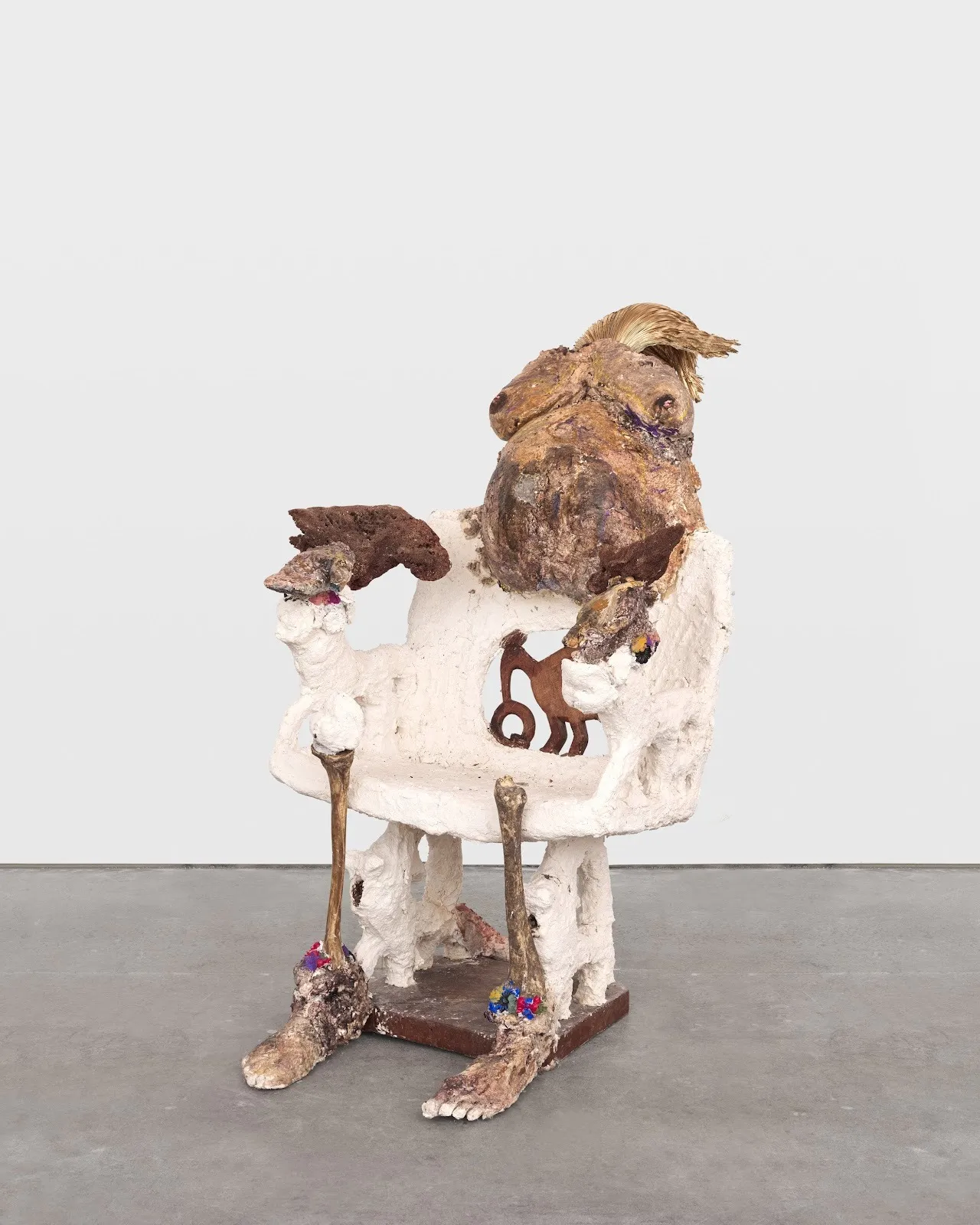 A lumpy white chair is affixed with two legs made out of bone and human feet at the front. The chair back is affixed with what appears as a pregnant torso with a heavily textured brown surface.