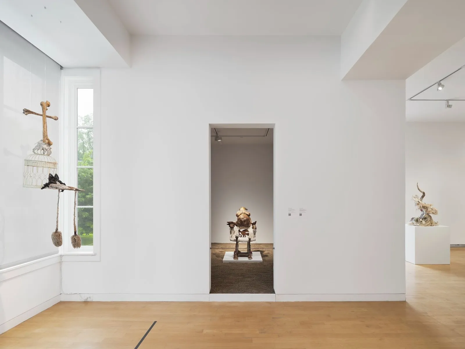 A gallery room with an adjoining smaller room entrance hosts a number of sculptural works, one of which appears as a bone cross. Through the doorway of the smaller room, a central sculpture appears as a chair-body hybrid.