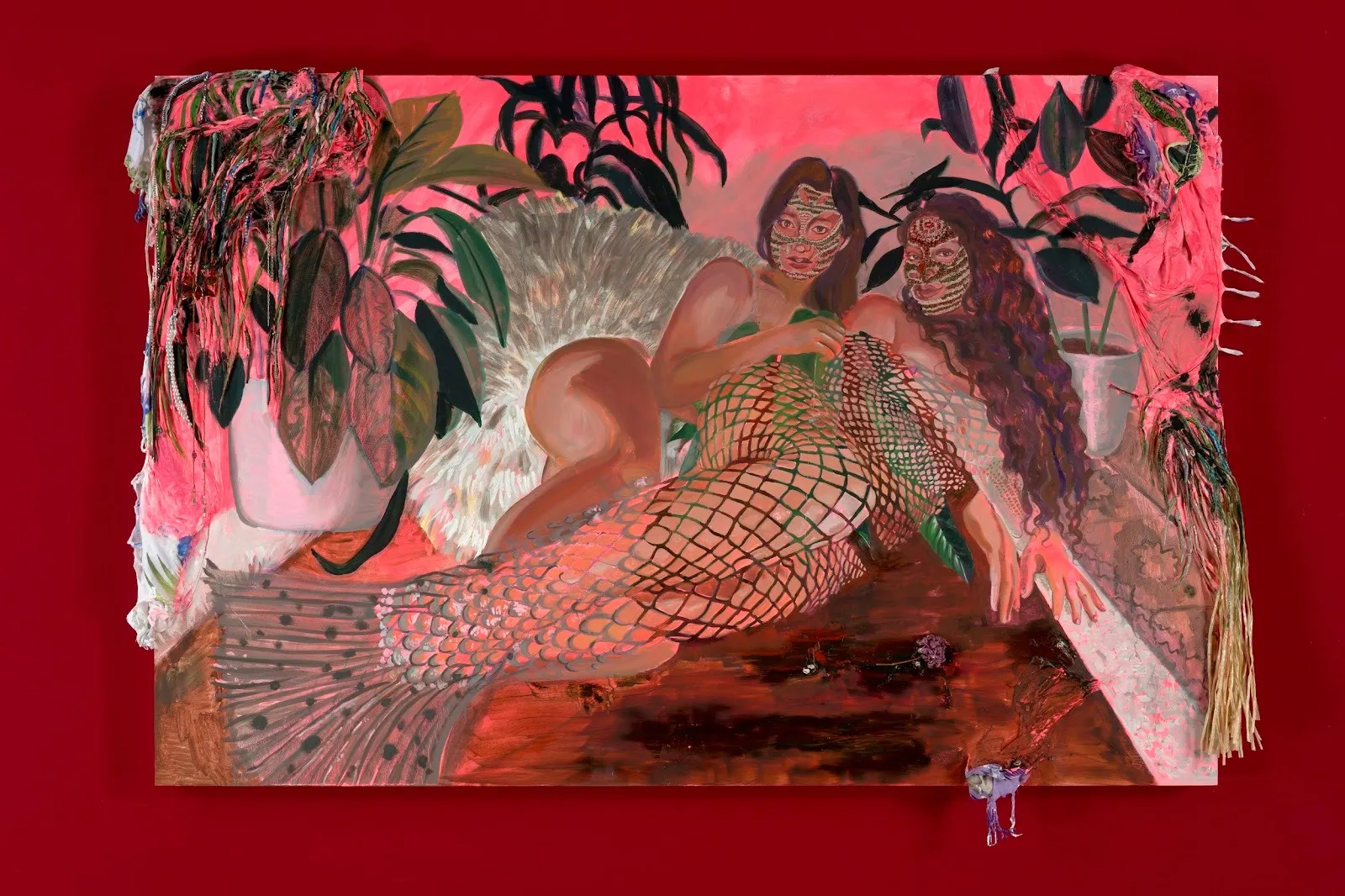 Against a red wall, a multicolored paintings features two people, one of whom has a mermaid-like tail, embrace each other in repose. Their faces are painted with mask-like designs. They are surrounded by potted plants and foliage.