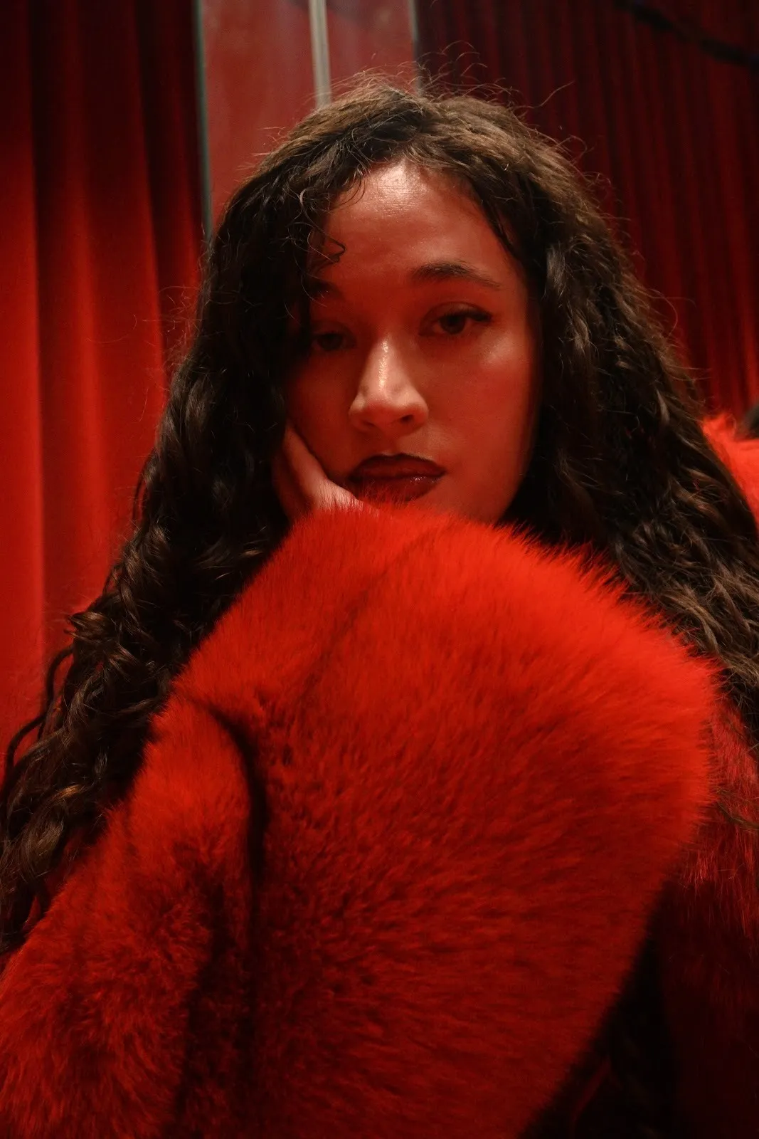 A woman with long, wavy hair wearing a red fur coat rests her chin in her hand and stares directly at the camera.