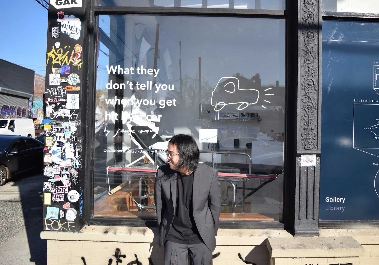 A man wearing a grey suit leans on the outer facade of a gallery-like space, looking to the left towards the side street.