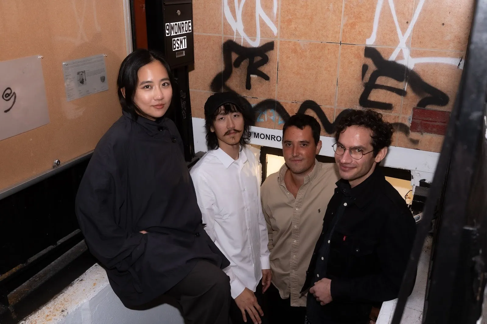 Four people stand grouped in a graffitied stairwell, looking at the camera and smiling.
