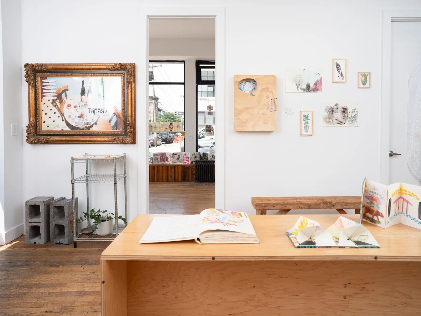 A white-walled space features multiple artworks on its walls and floors. Foregrounded is a raw wooden table with colorful ephemera and open books.