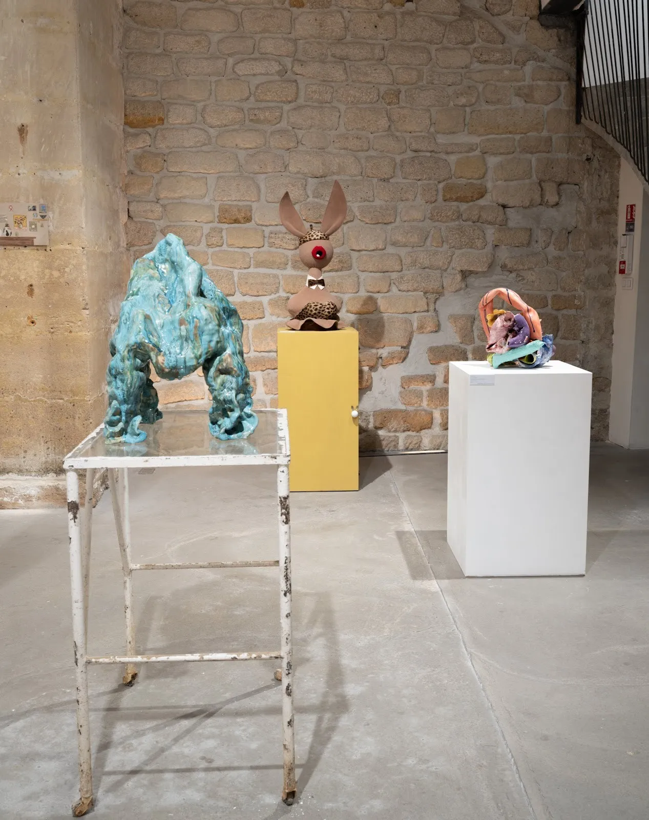 Three colorful abstract sculptures displayed on separate pedestals against a stone wall in a gallery.