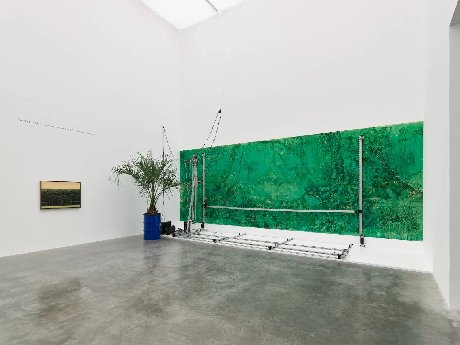 In a white gallery room, a small painting of a forest hangs on one wall. On the other, a large, green painting of a forest is hung to a wall. Attached to it are metal bars and wires on its surface and on the floor. A palm tree in a metal barrel sits.