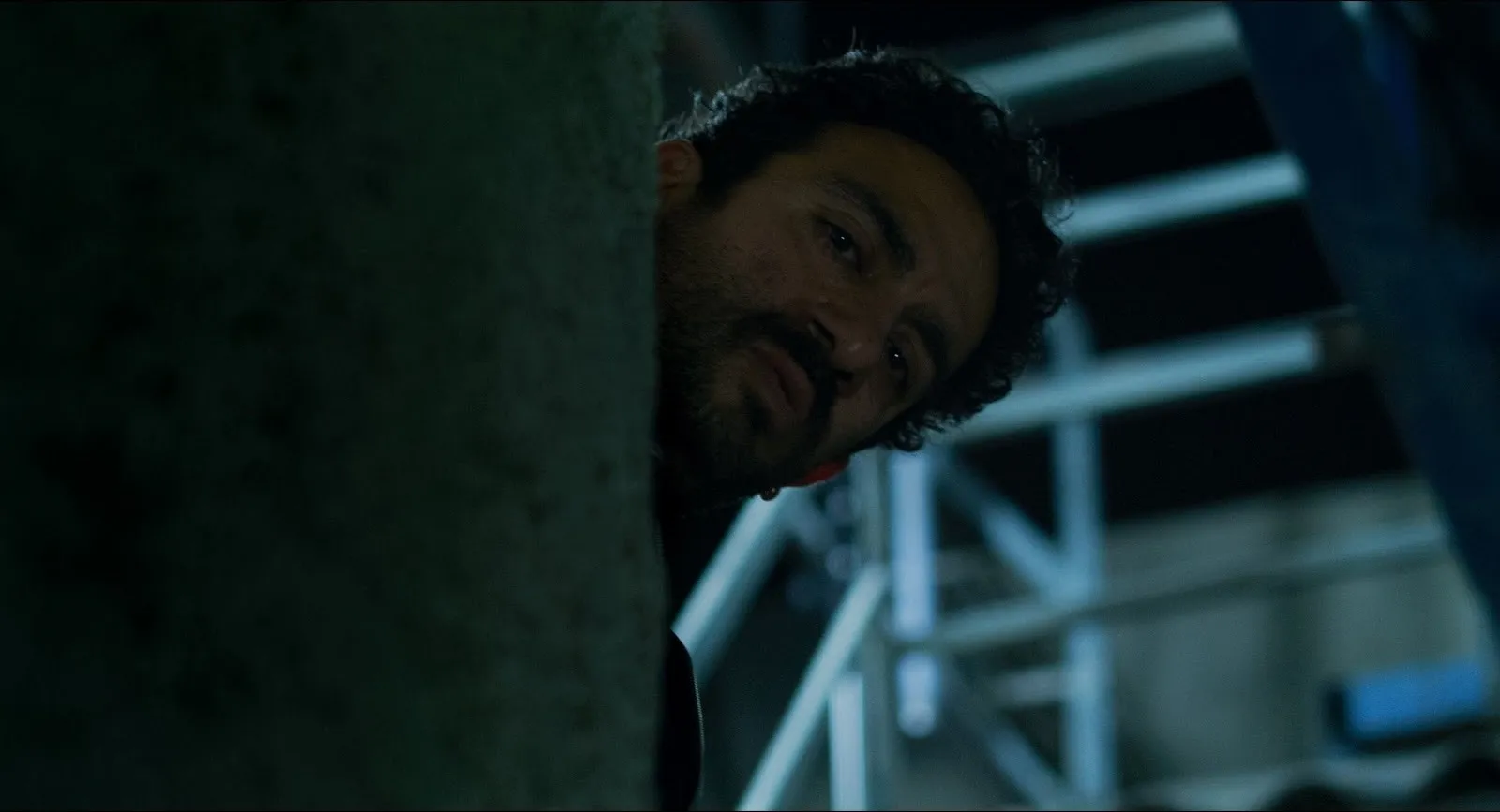In a dimly lit building with stairs behind, a man looks around a corner, only his head visible.