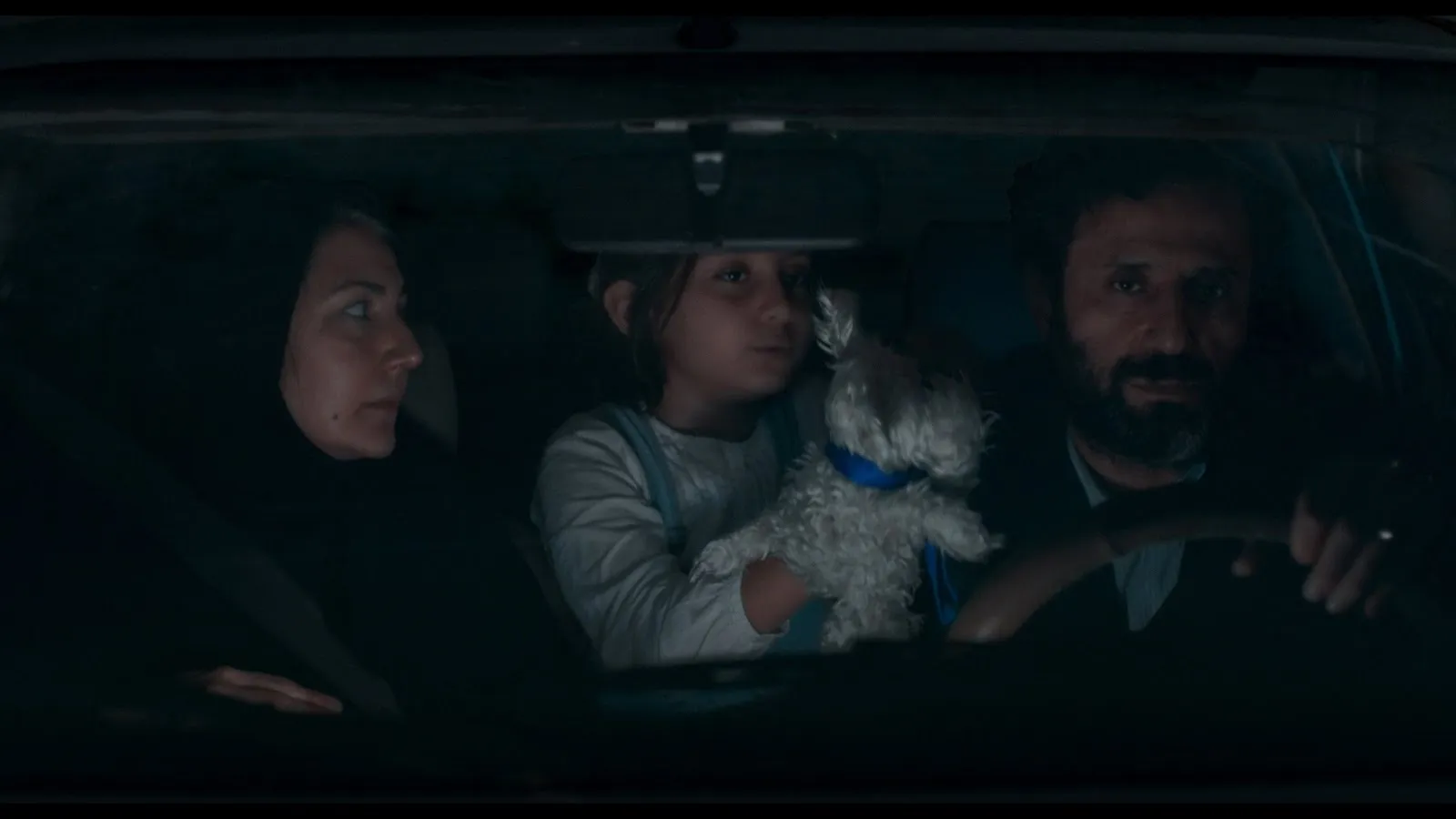 In a dimly lit car at night, a man drives somberly, while the woman in the passenger seat looks at him. A child in the middle holds a white plush stuffed animal.