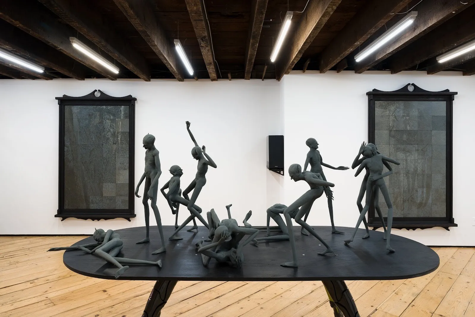 In a white gallery room with a wooden beamed-ceiling, a large, central, black metal table supports a variety of black metal humanoid figures in various states of movement. On the far wall, ornate wood-framed pieces are made of grey panels in grids.