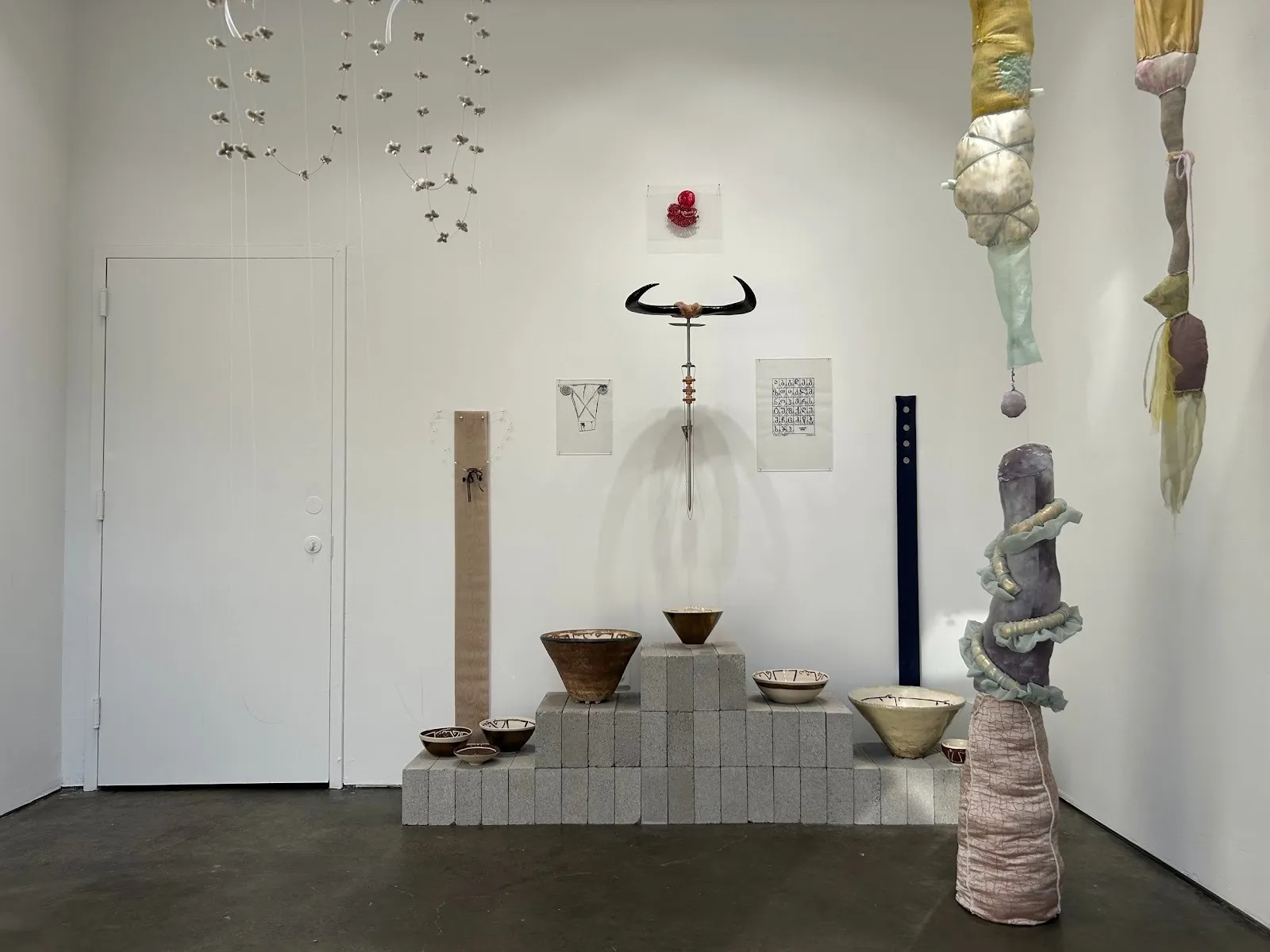 A white gallery room is populated with various sculptures, both hanging and fixed. Such sculptures seen are bowls, delicate wire structures, and lumpy, semi-organic fabric columns.