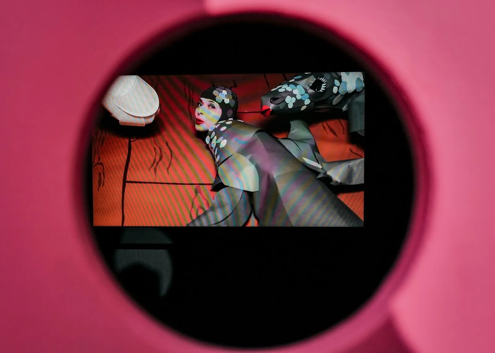 Through a pink pinhole, a screen can be seen. On the screen, a woman clad in structural grey clothing against an orange background looks directly at the camera, mid-motion.