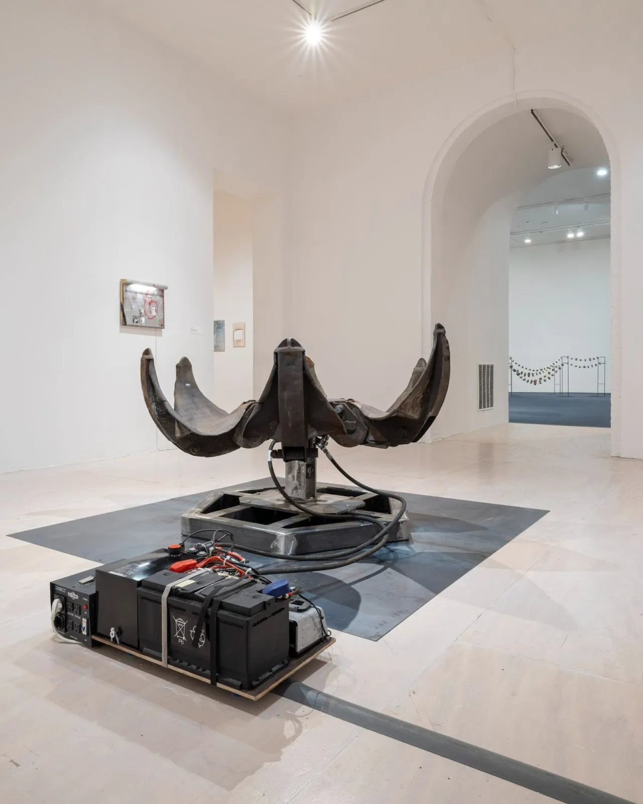 In a white room, a central sculpture is made of an electric motor attached to wires and a large metal six-petal flower-like sculpture.