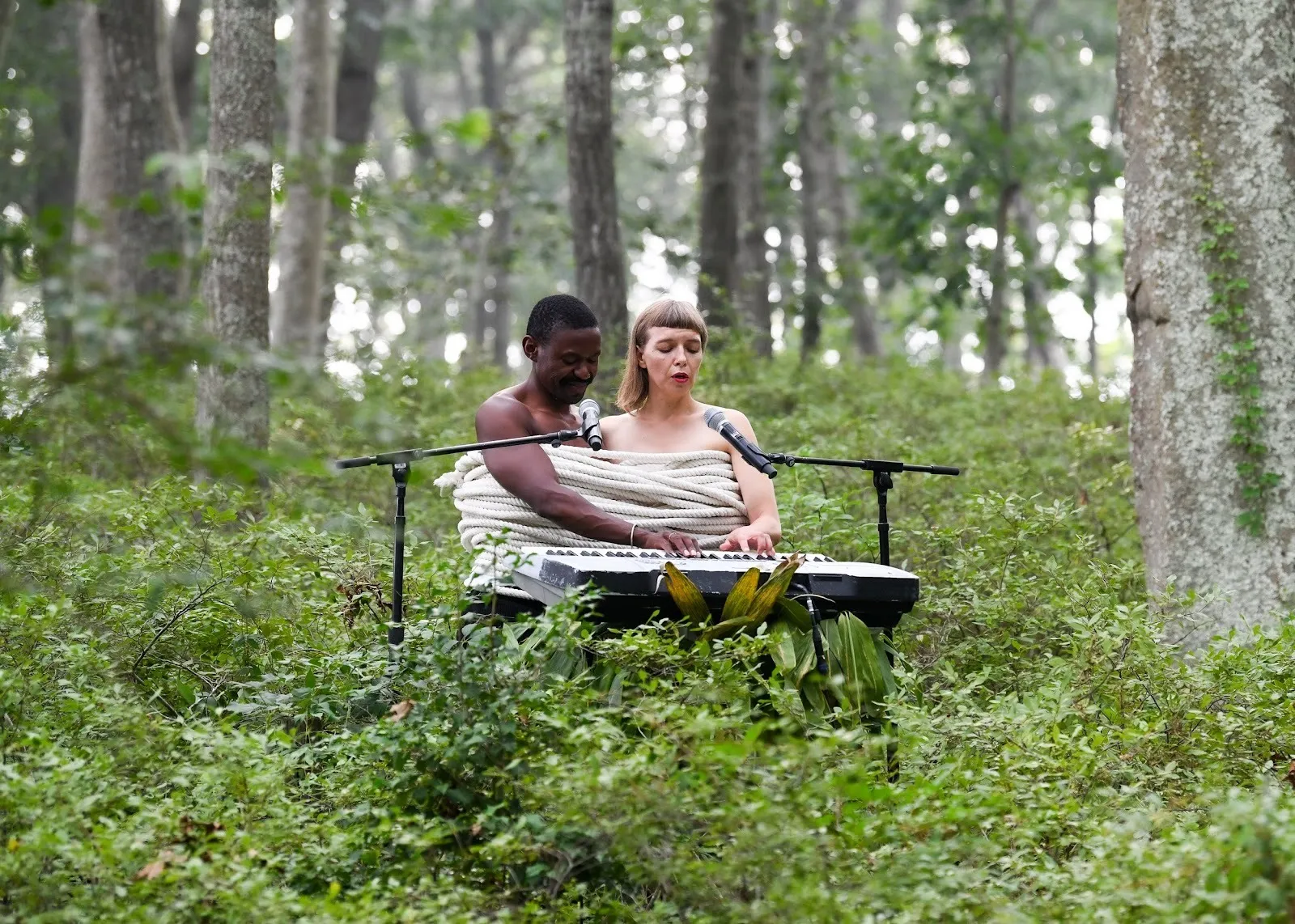 In a foliaged forest setting, two people sit, torsos tied together by thick white rope, and perform on one keyboard and two microphones. Each performer has one hand free which plays on the keyboard.