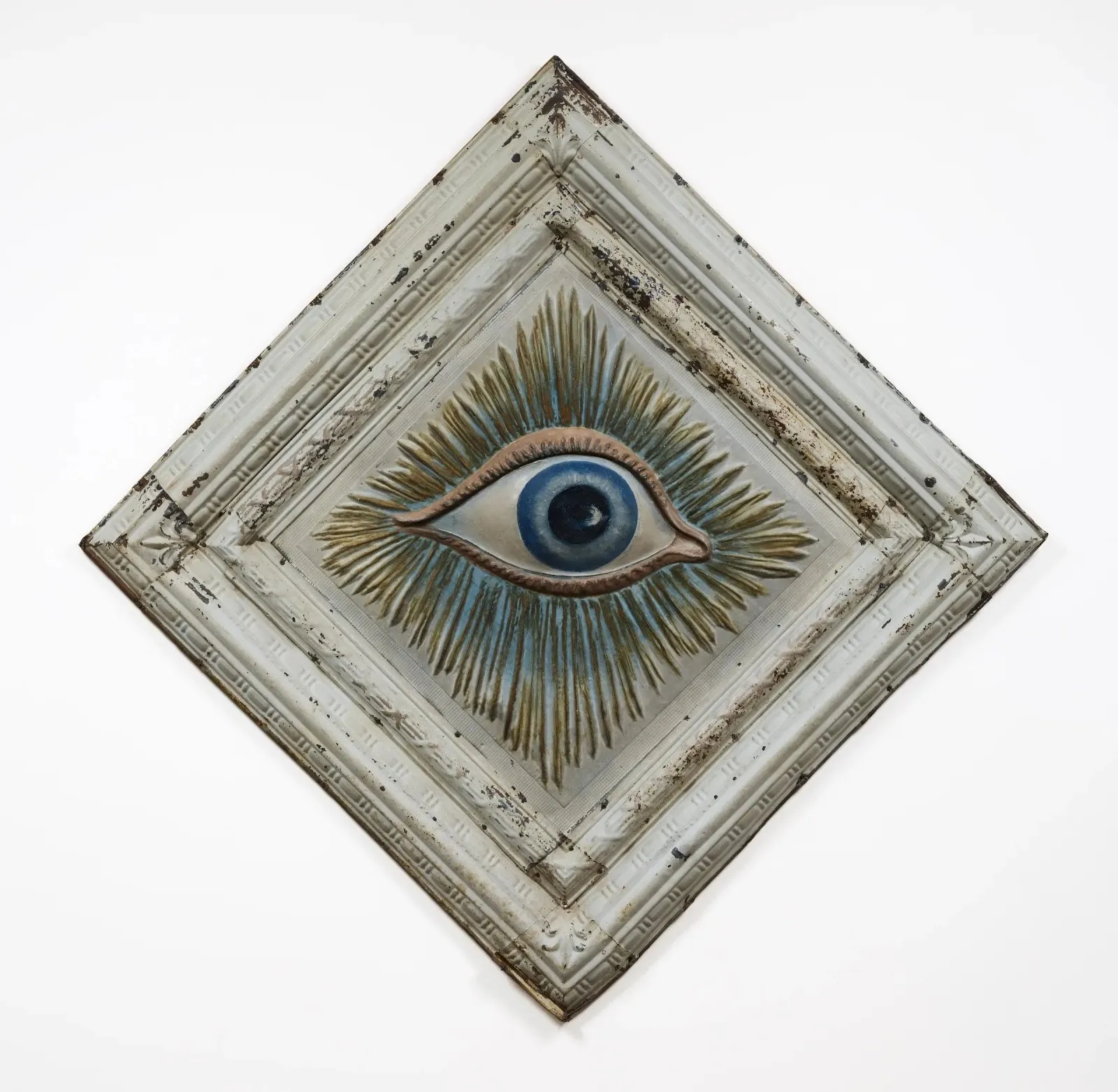 A worn-down white square frame, configured in a diamond shape, frames a painted relief of an open blue eye, surrounded by line-bursts of blue and gold.