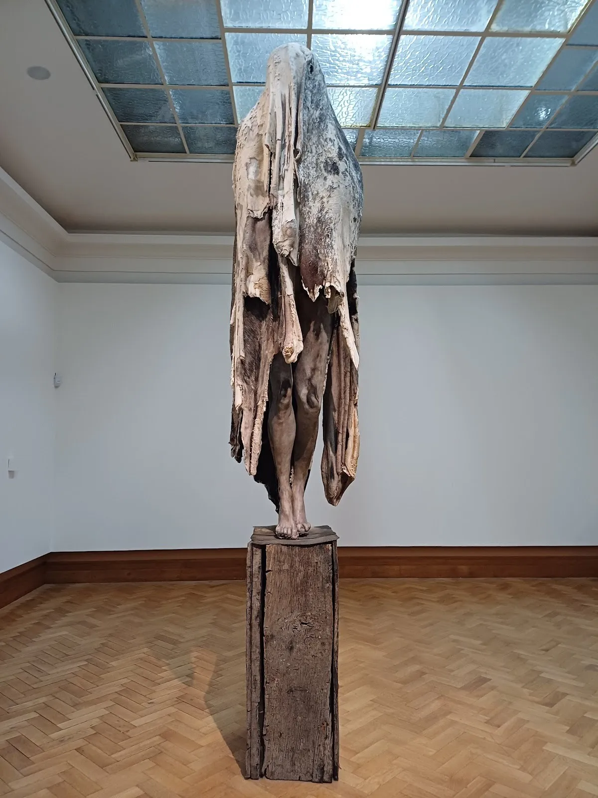On a tall, skinny rough wood plinth, a human-like sculpture is draped with worn, white and brown cloth, obscuring its form but revealing the figure's legs, which seem to be in light mid-step.