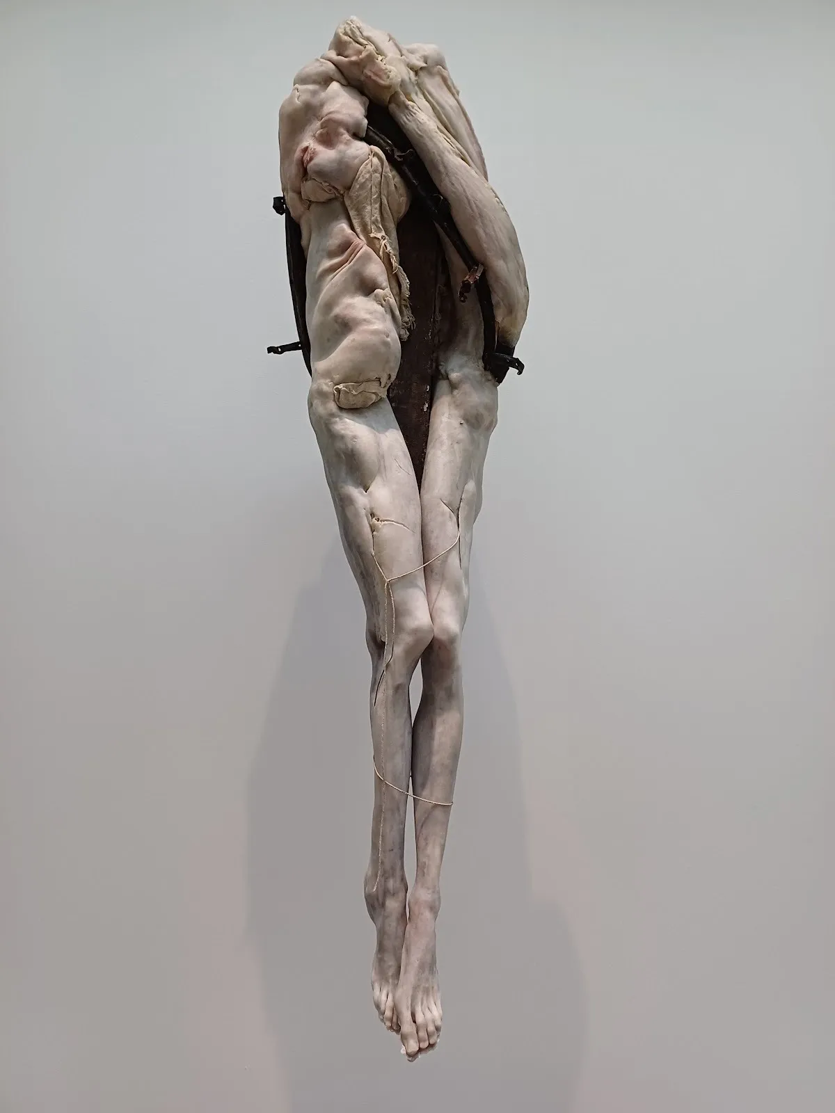 A pale, humanoid figure with long legs and a distorted, fleshy top is affixed to a metal structure and mounted to a wall. The legs are loosely bound by thin string.