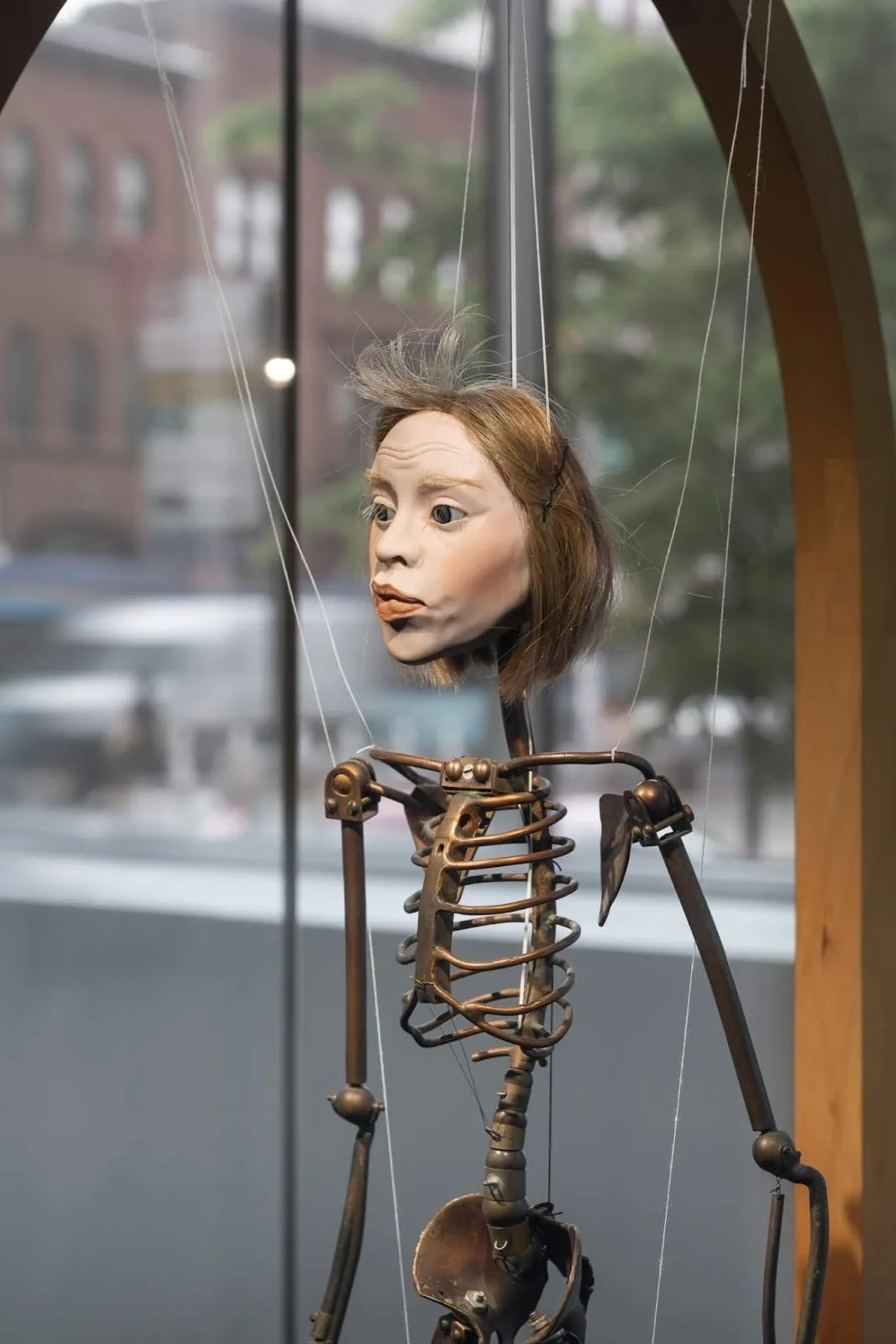 Sculpture of a doll head affixed to a metal, hybrid skeleton-machine body. The whole figure is held up by thin strings tied to various joints on the figure.