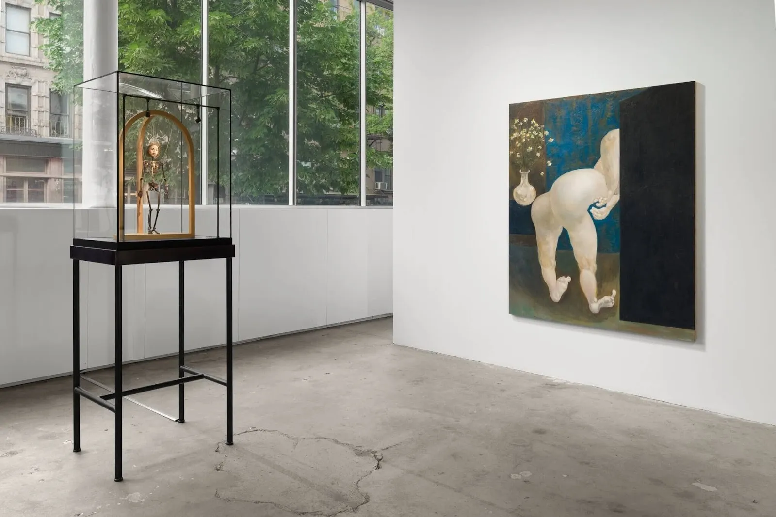 In a white gallery room with windows facing a street, a box vitrine on a table features a skeletal doll-like work. On the right wall, a mid-sized painting features an abstracted body next to a flower vase; the bent figure's torso is obscured.