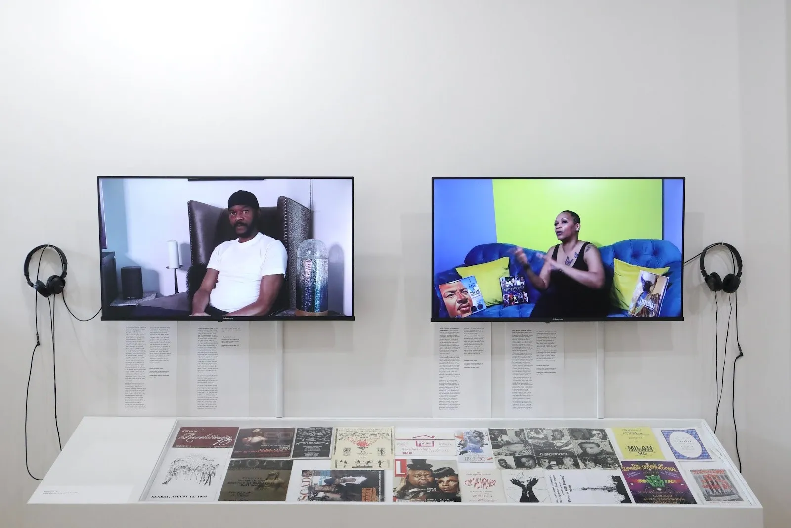 Two monitors are mounted side-by-side on a white wall with headphones attached. Each monitor displays a subject being interviewed. Below, a flat vitrine holds various paper ephemera.