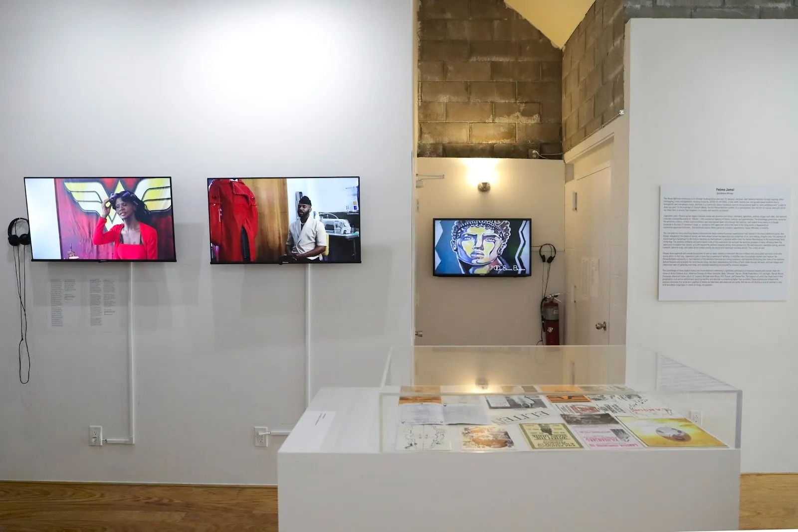 A white gallery room holds multiple works. On the left, two monitors play. In the center, a vitrine showcases paper ephemera. In the back, another monitor plays.
