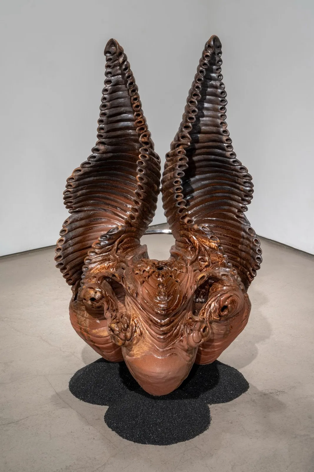 A sculpture, placed atop a pile of black sand on the floor of a white gallery room, takes an abstracted form resembling a horned animal. Suggestions of a face and horns are made of detailed and almost symmetrical configurations and layers of clay.