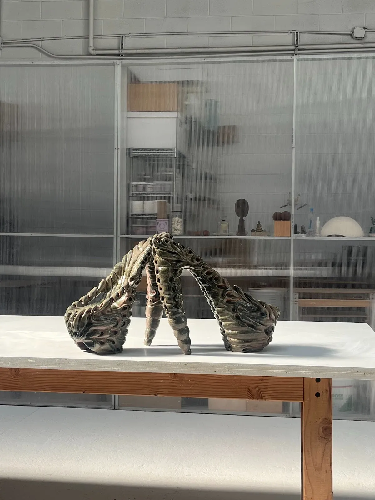 Two forms resembling platform stiletto shoes sit atop a table. The shoes are designed from bulbous, liquidy layers of clay.