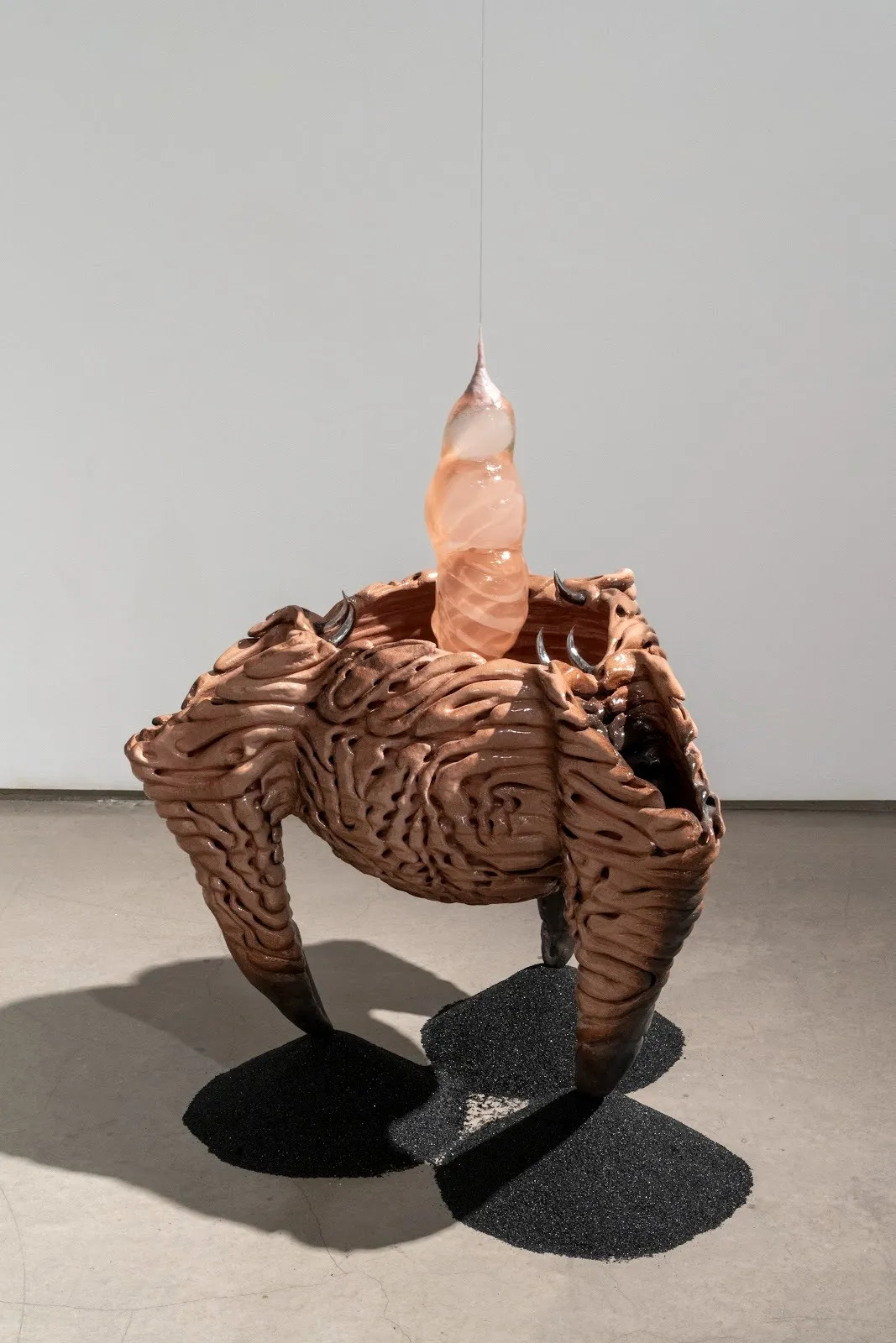 A tripod-cauldron, sitting atop three mounds of black sand, is made from layers of patterned clay. A light orange glass piece, shaped like a chrysalis, hangs from the ceiling into the cauldron.