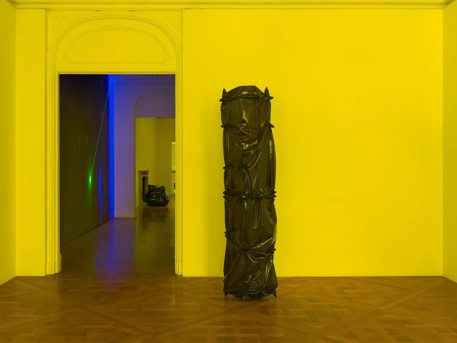 A yellow-lit room with hardwood floors features once tall sculpture made of wrapped and pleated latex, standing in a column. Through an open doorway, blue and green lighting washes another room.
