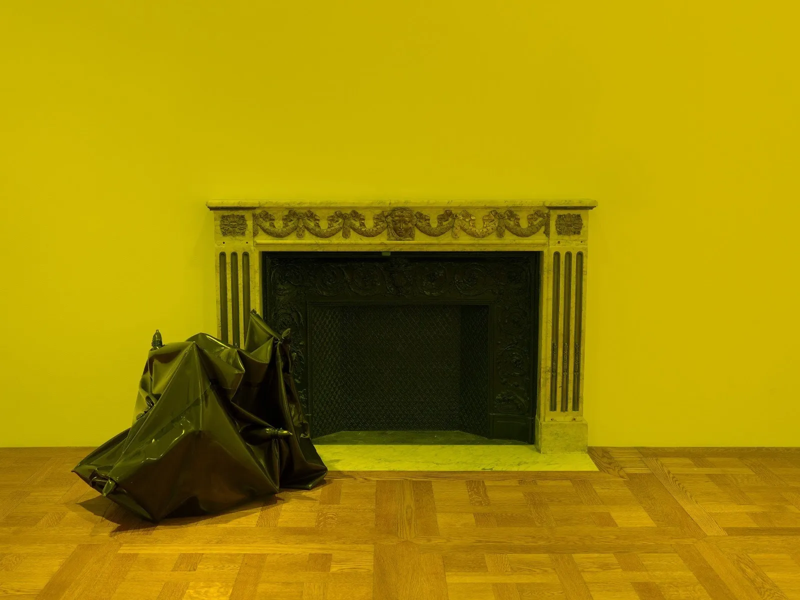 In a yellow-lit room with hardwood floors, a medium-sized latex structure, similar to a trash bag, rests next to a fireplace.