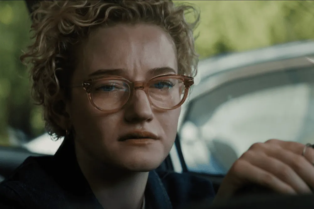 A woman with short, blonde, curly hair and glasses sits inside of a car and seems to be near tears, or actively crying.