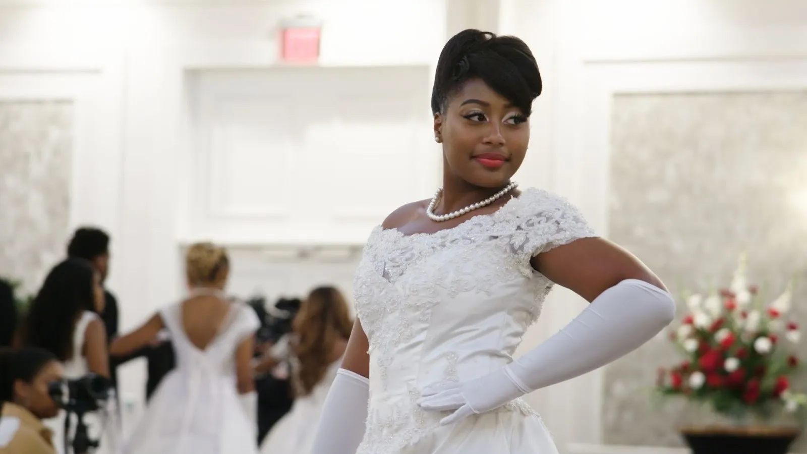 A woman poses as if looking into a mirror off-camera, wears a white debutante dress, opera-length white gloves, and a pearl necklace. Other debutantes mill about in the background.