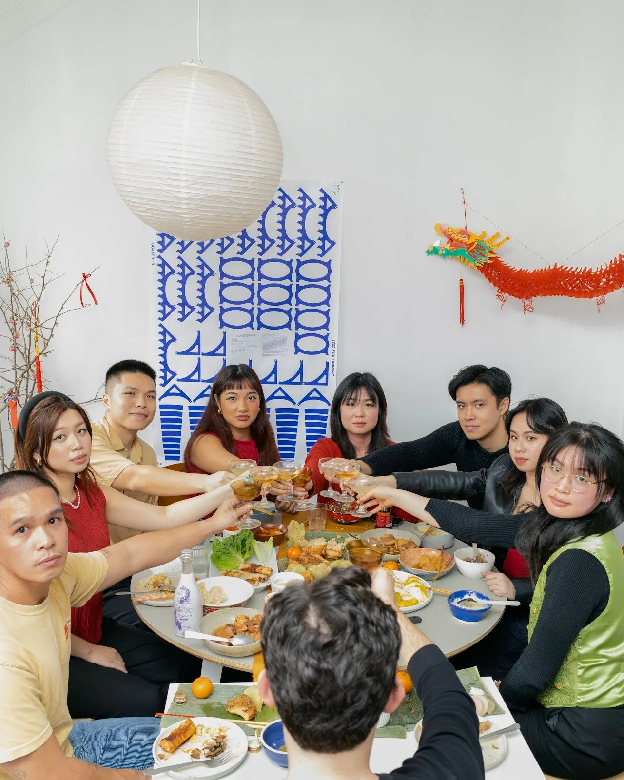 In a room decorated with posters and paraphernalia, nine people sit around a food-laden table and hold their drinks up, mid-cheer, while staring at the camera.