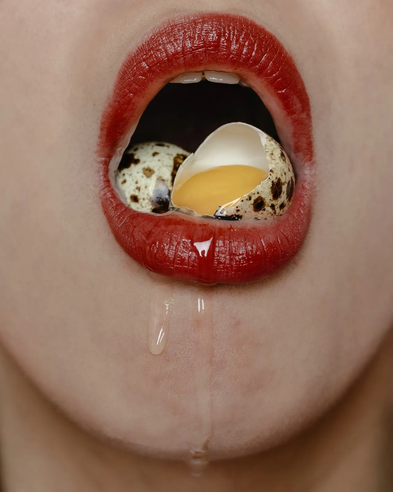A close-up shot of lipstick-painted lips open to reveal a crushed quail egg. The egg yolk sits inside of the crushed shell, while the clear egg white runs down the person's chin.