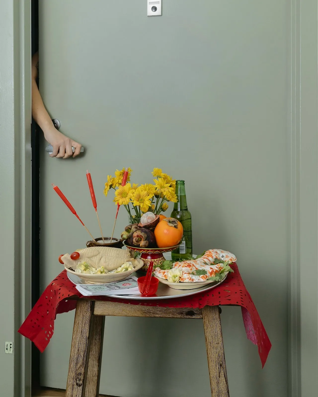 Against a grey door held slightly ajar by a hand gripping the handle from the inside, a stool covered with a red tablecloth holds a platter of food, a stack of money, a beer bottle, a bowl of fruits and flowers, and a pot holding incense sticks.