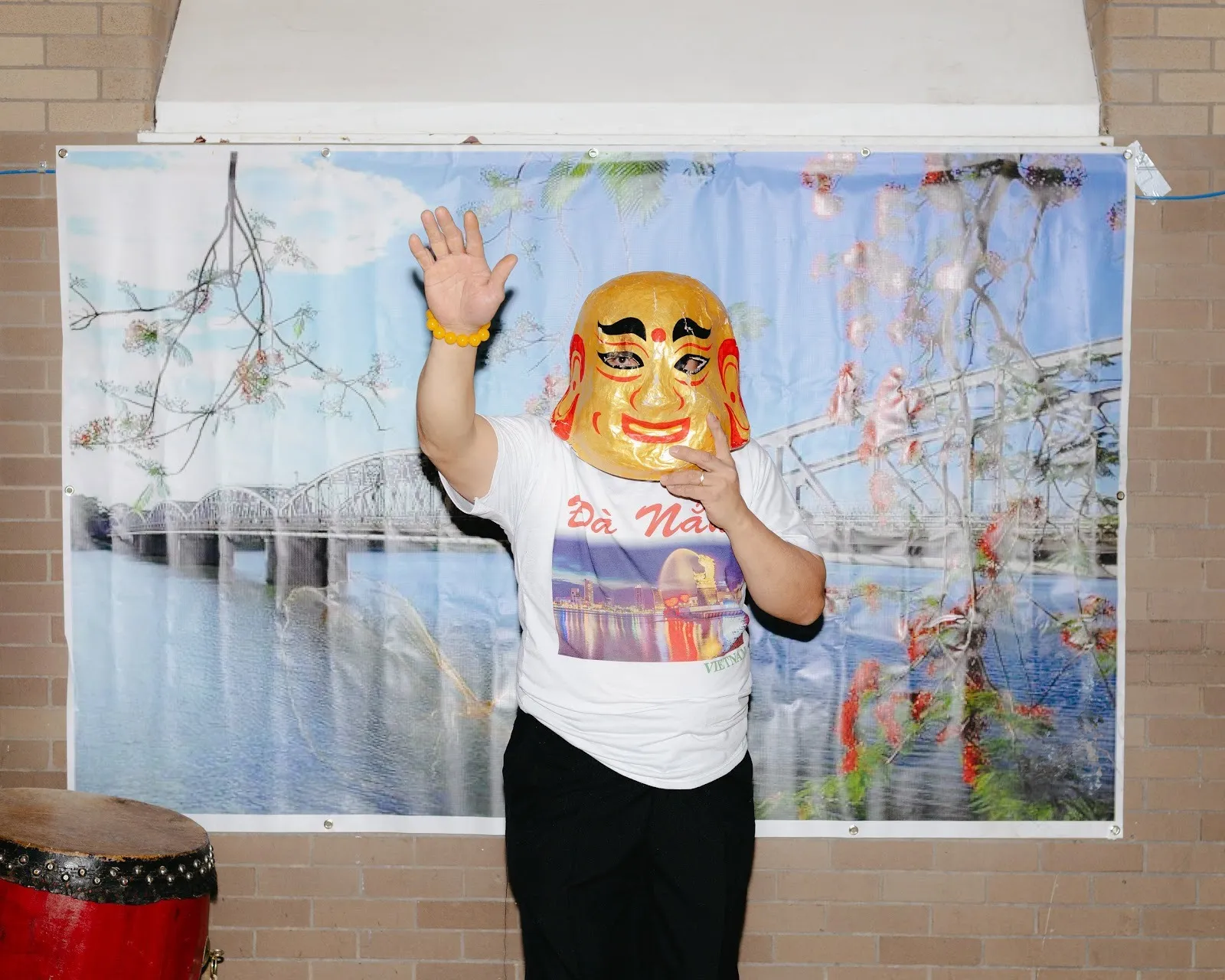 Against a light brick wall with a poster of a landscape pinned to it, a person holding a golden, smiling mask to their face waves to the camera.