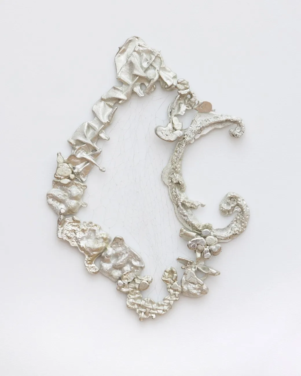 Mounted to a white wall, an ornamental, silvery pewter frame is made from detailed, worked metal and features organic, curling designs. The frame forms a curved, oblong outline. Inside the frame, white wall/plaster is filled with tiny cracks.