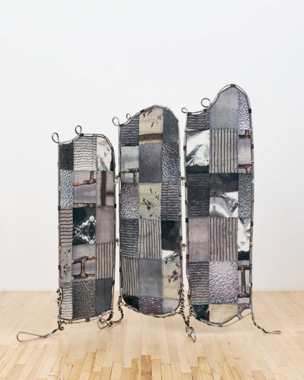A folding screen stands in a white gallery room. The screen is made from thick, twisted wire, and the stretched fabric is made from a patchwork of different grey textiles, some patterned, some with images printed on.