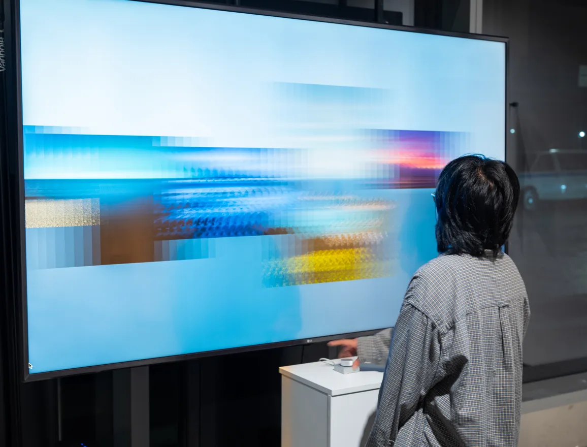 A person manipulates a small button on a plinth, looking up at a large tv monitor which displays a colorful, pixellated blur.