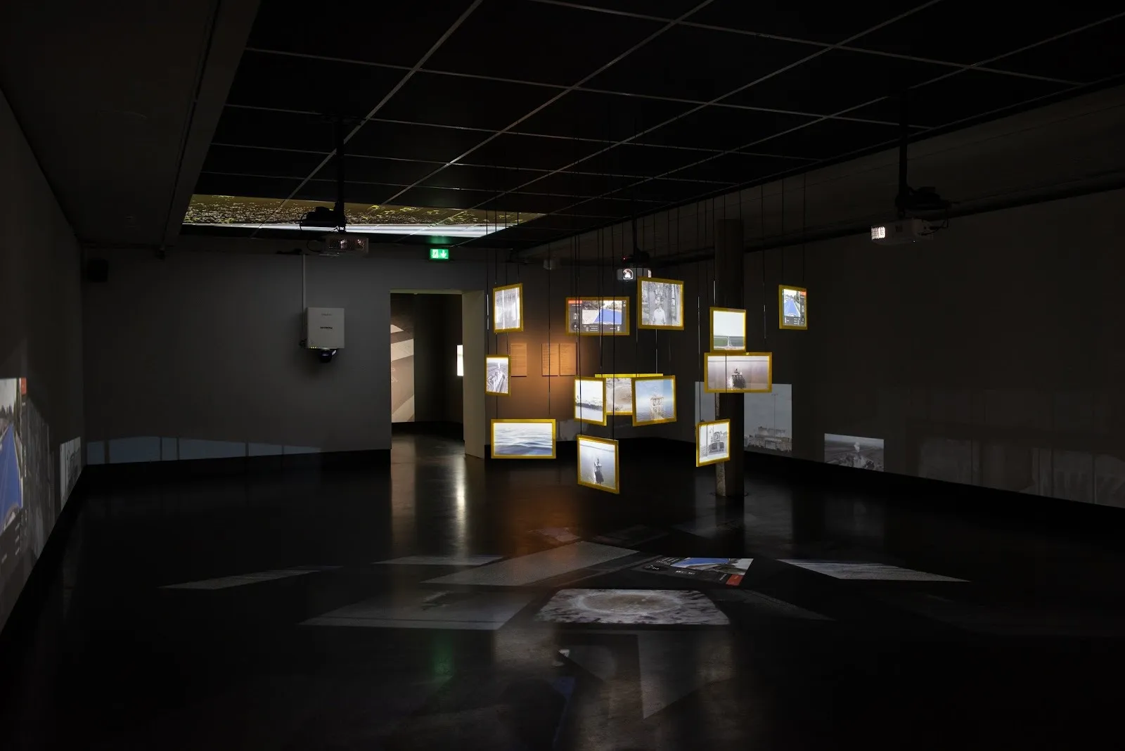 A dark gallery room features a central installation of hanging illuminated framed pictures. They hang scattered at different lengths, their projected silhouettes appearing on the floor.