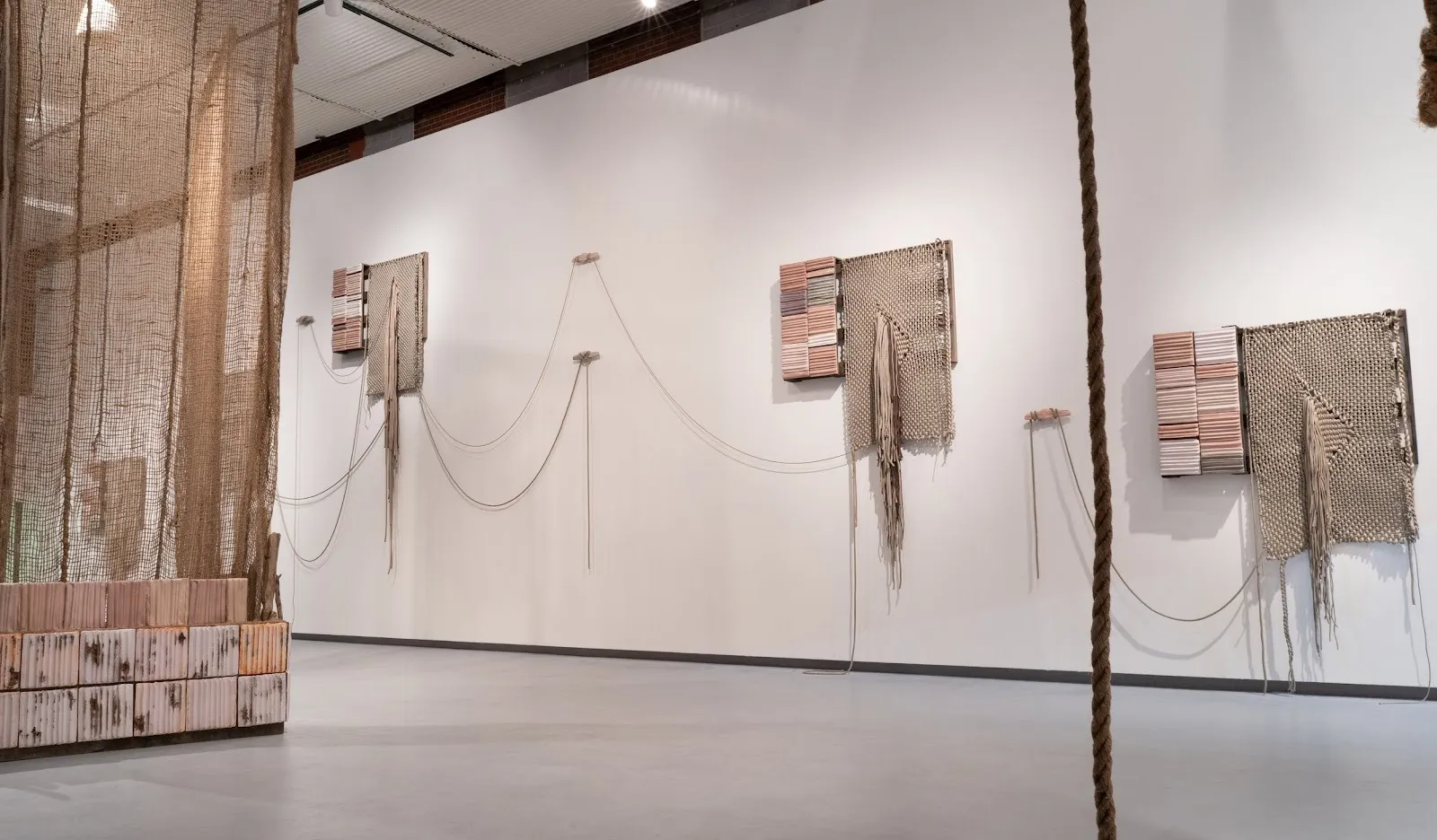 Large, beige textiles hang from a large white gallery wall. Another textile piece hangs from the ceiling, translucent and beige.