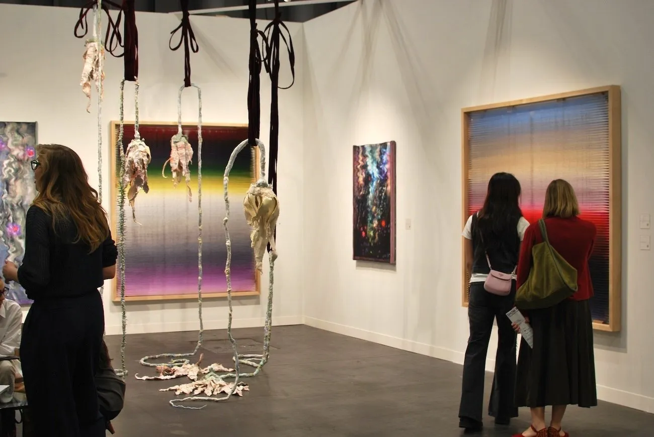 In a white gallery booth, several 2-D and 3-D works populate the space. The paintings are colorful, featuring wavy hue gradients and brushstrokes. The sculptures are fabric-like tendrils that hang from the ceiling.