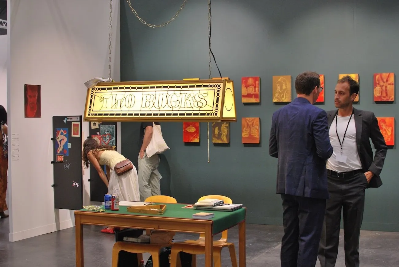 In the corner of a gallery booth with one white and one grey wall, several small orange and beige works hang in a grid. In the center of the floorspace, a table with beer cans stands under a long bar lamp with glass that reads "TWO BUCKS."