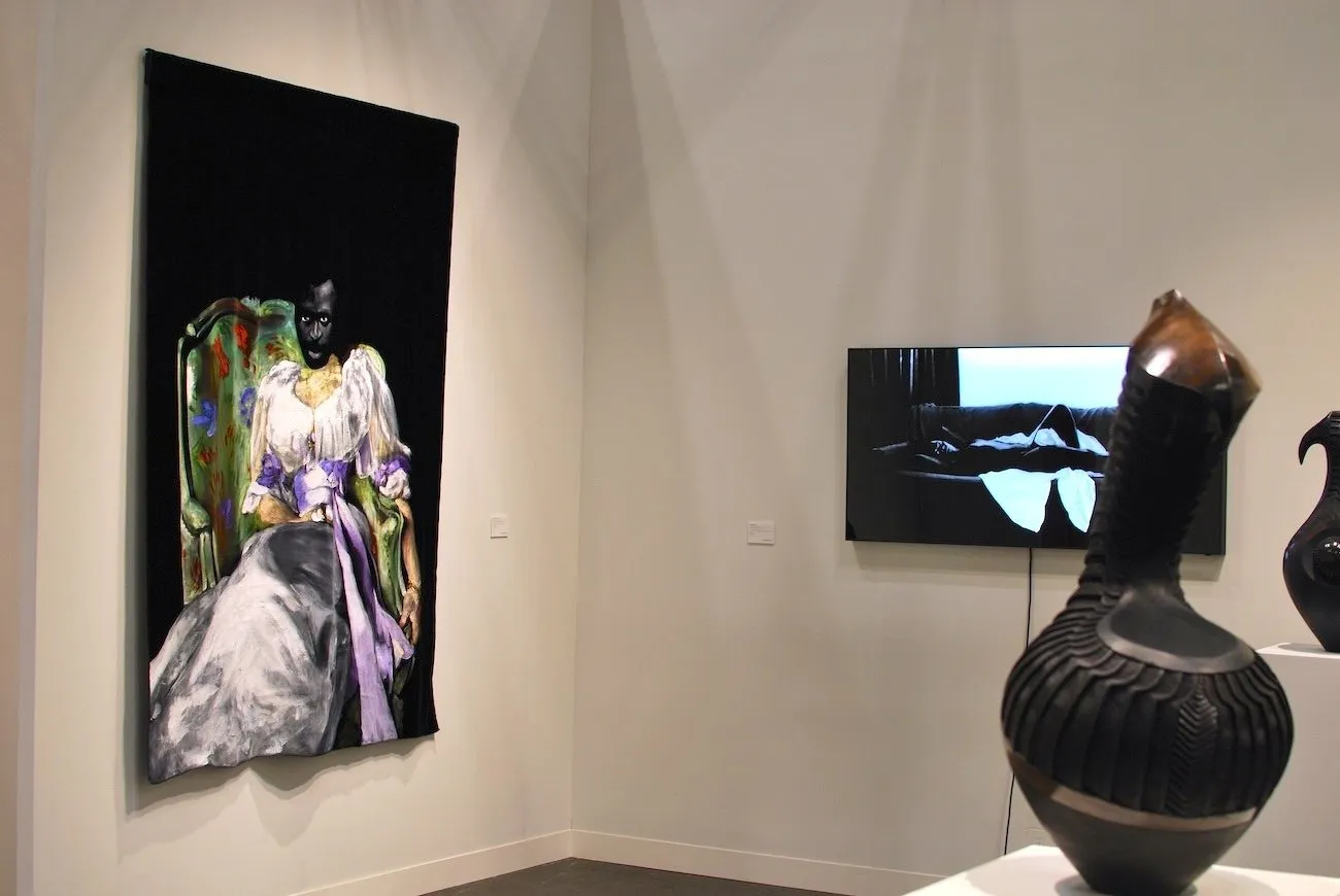 In a white gallery booth, a painting, a monitor, and a gourd-like sculpture on a plinth populate the space. the painting is a portrait of a seated figure in an elaborate dress.