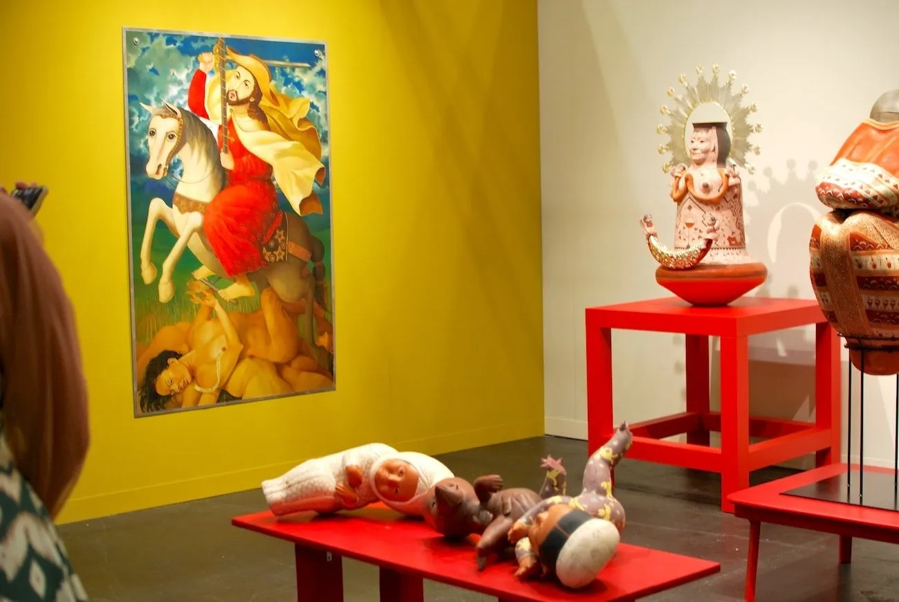A gallery booth with one white and one yellow wall features various sculptural and 2-D works. The painting is a stylized, abstracted rendition of a Catholic saint depiction. The sculptures rested on red tables range from baby dolls to totemic idols.