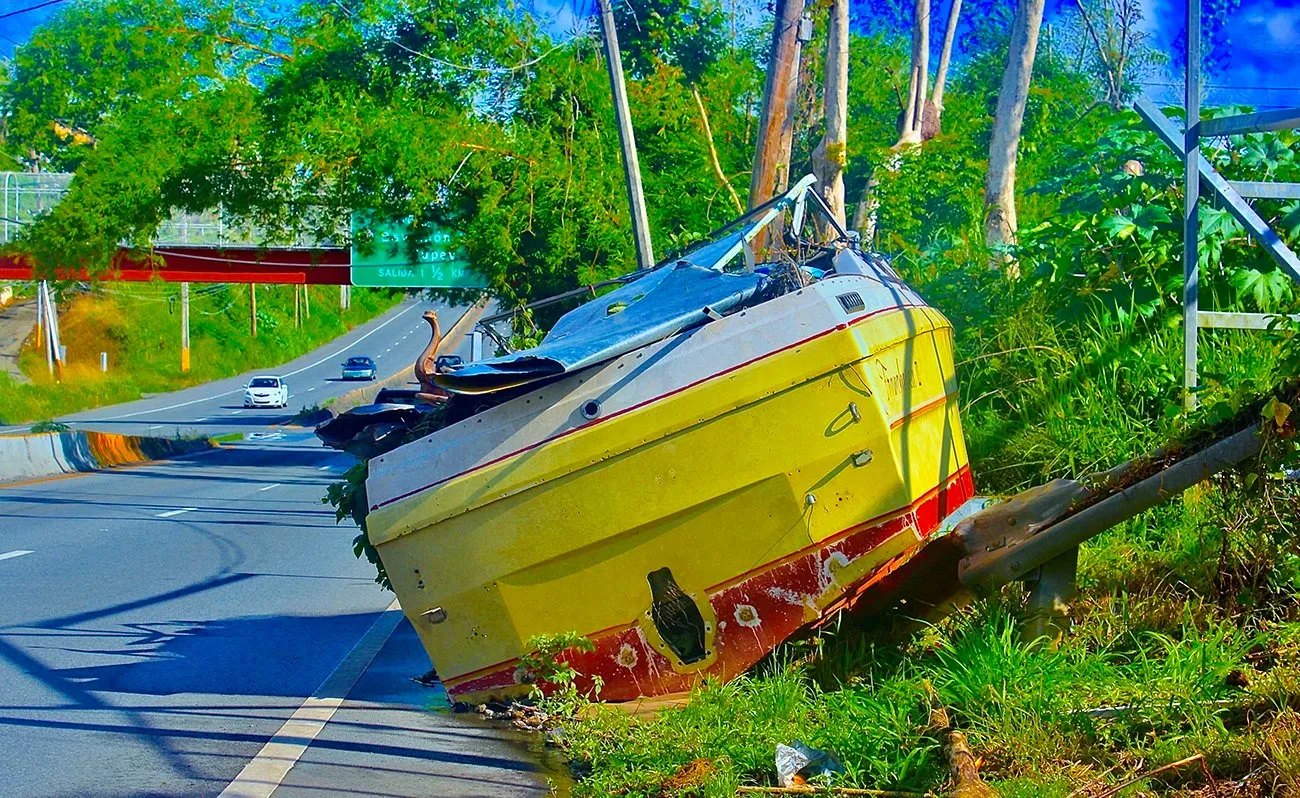 On the side of a highway, a beached yellow boat with a red bottom leans half on asphalt and half on foliage and structure.