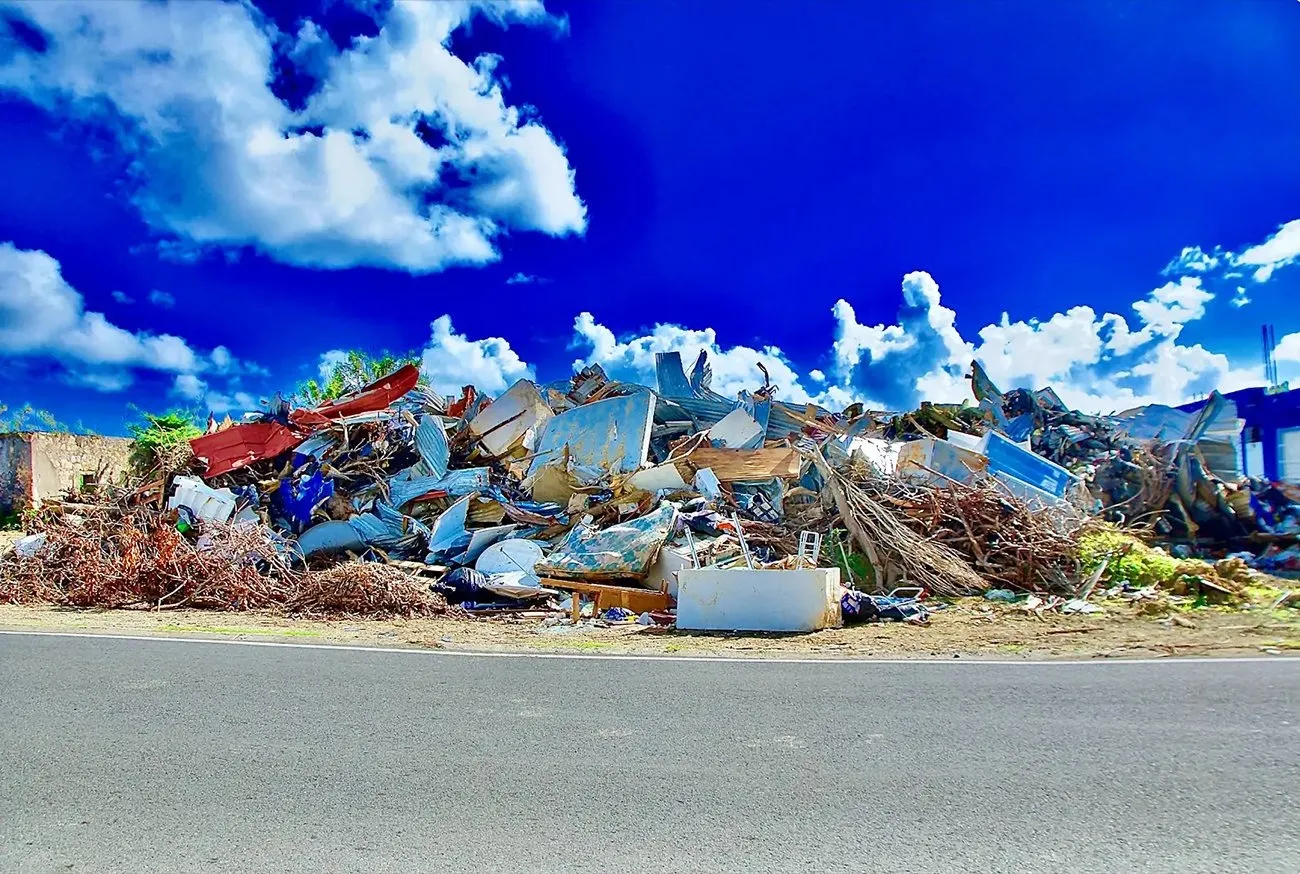 A large pile of detritus—concrete, metal, brush of different colors—stand on the side of a road against a bright blue sky with clouds.